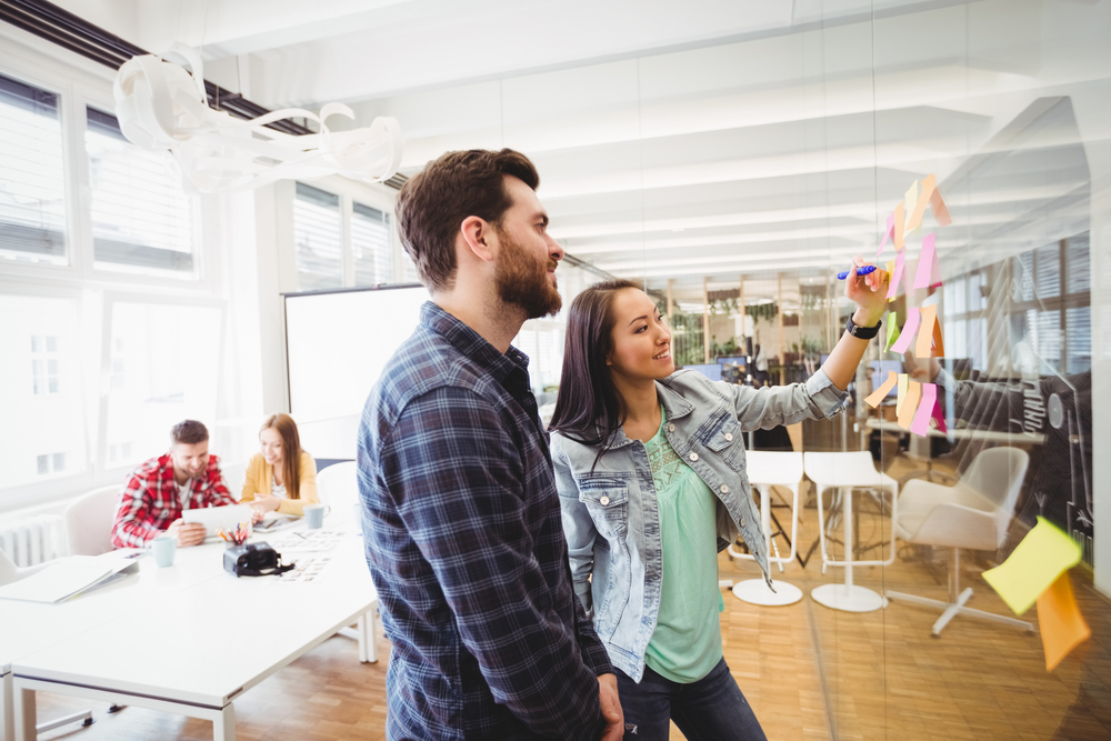 Smiling business people looking at sticky notes on glass in meeting room at creative office